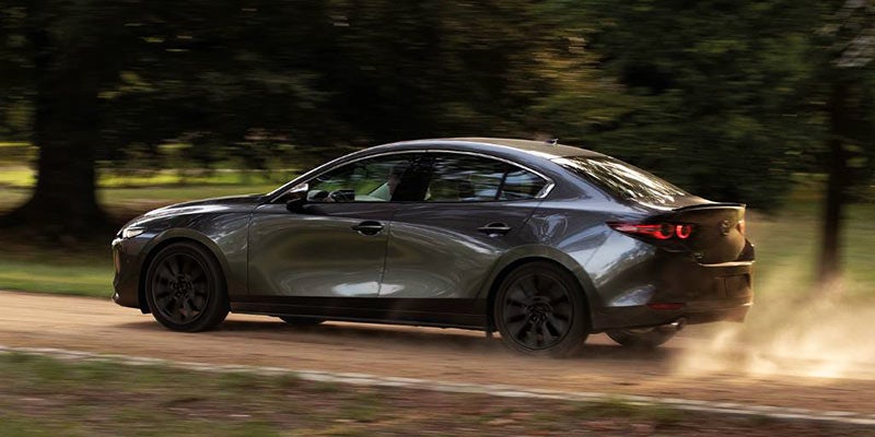 Dark gray sedan driving on a dirt road, kicking up dust at the rear, with trees in the background.