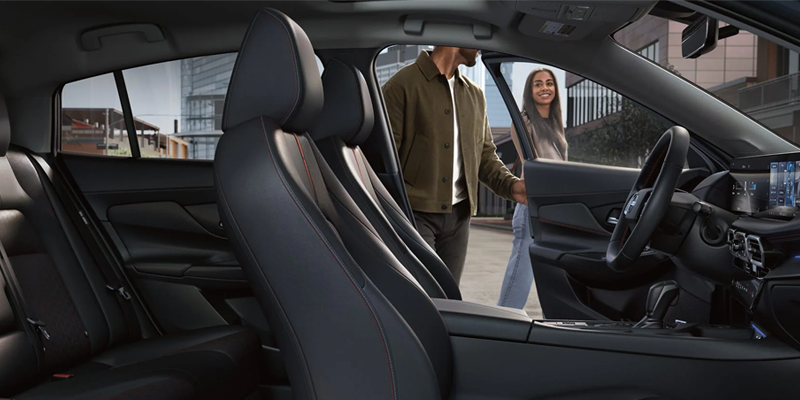 View from inside a car: a man and woman enter the car through an open front passenger door, smiling.